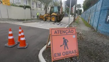 Imagem mostra uma rua bloqueada com uma placa de obras e uma máquina na pista.