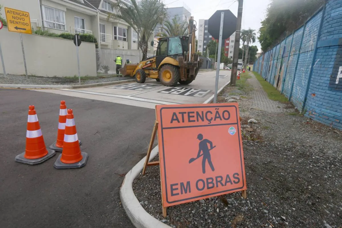 Imagem mostra uma rua bloqueada com uma placa de obras e uma máquina na pista.