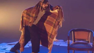 Imagem mostra um homem em cena. Ele está com uma coberta nas costas e ao lado de uma cadeira.