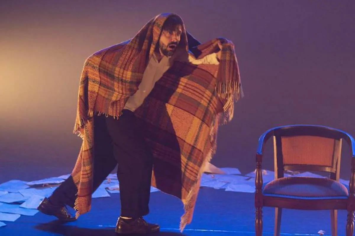 Imagem mostra um homem em cena. Ele está com uma coberta nas costas e ao lado de uma cadeira.