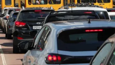 Imagem mostra carros em um congestionamento em Curitiba. No destaque, um carro branco parado em uma rua com sol e um ônibus amarelo ao fundo.