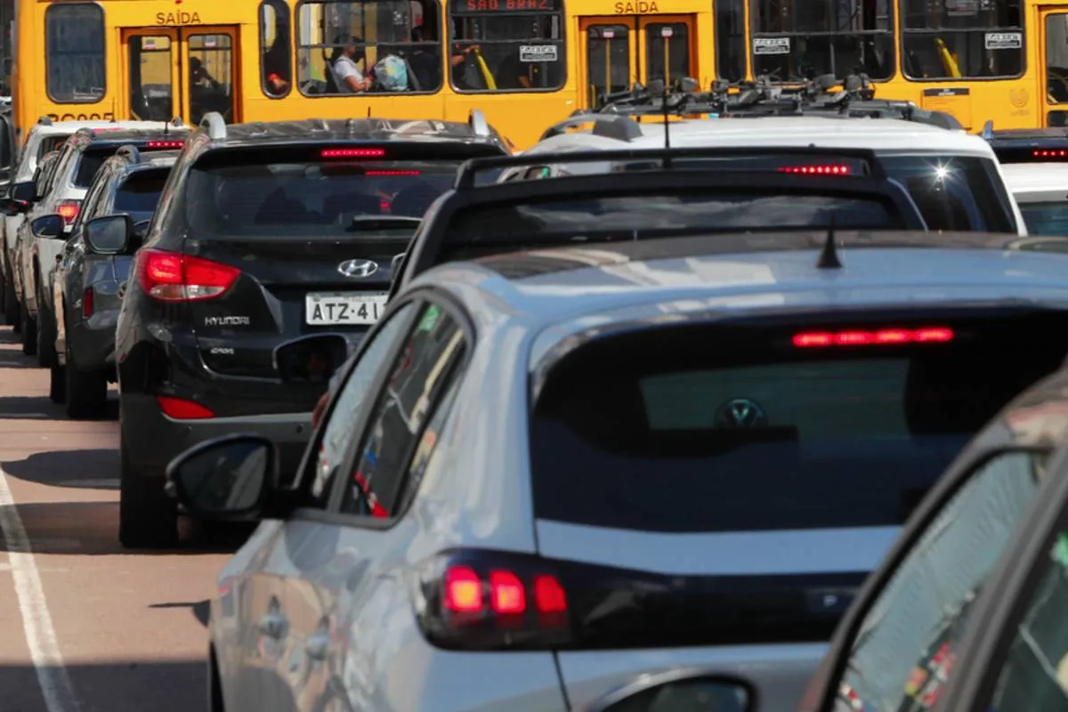 Imagem mostra carros em um congestionamento em Curitiba. No destaque, um carro branco parado em uma rua com sol e um ônibus amarelo ao fundo.