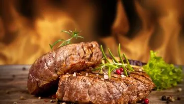 Imagem mostra dois steaks recém saídos da grelha, apoiados um no outro com labaredas da churrasqueira ao fundo. Os bifes estão em uma tábua de madeira com temperos e sal grosso.