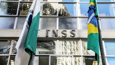 Imagem mostra a fachada de uma agência do INSS no Paraná, com uma bandeira do Paraná ao lado esquerdo e uma bandeira do Brasil do lado direito.
