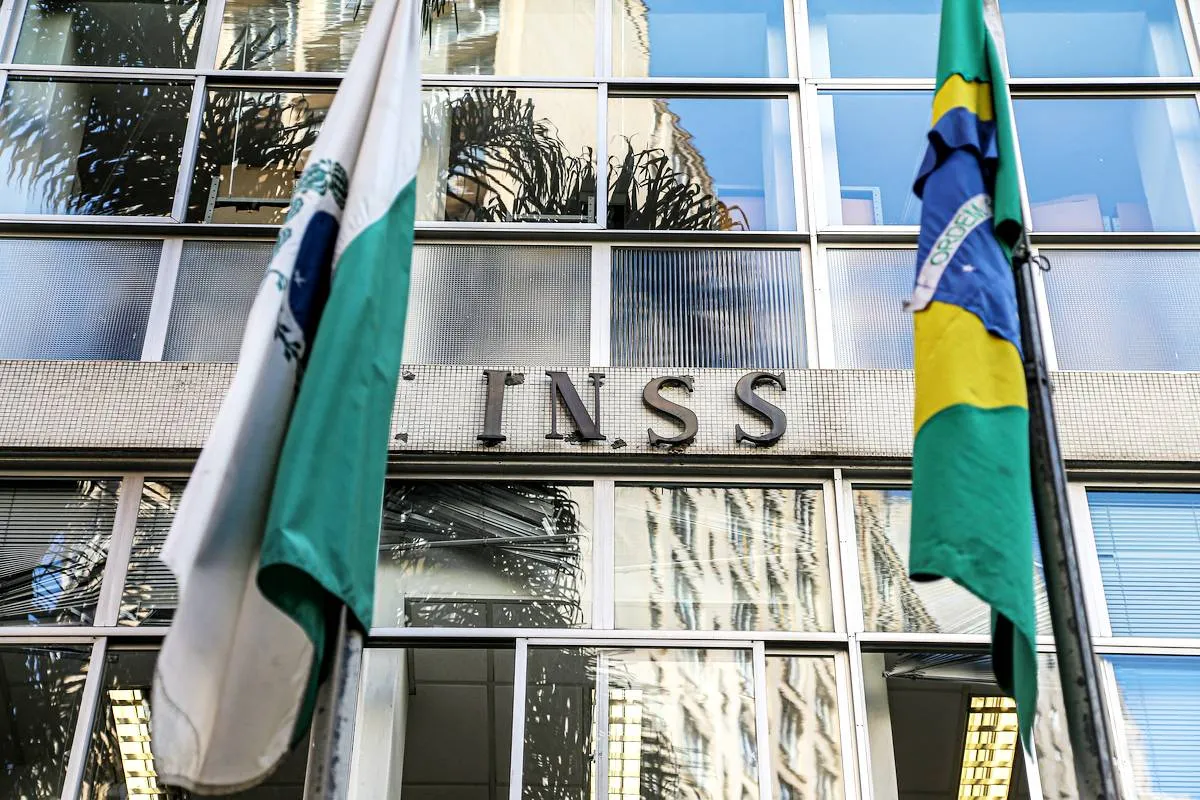 Imagem mostra a fachada de uma agência do INSS no Paraná, com uma bandeira do Paraná ao lado esquerdo e uma bandeira do Brasil do lado direito.
