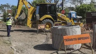 Imagem mostra uma obra no meio de uma rua de Curitiba. Há uma retroescavadeira e uma placa de 'trânsito impedido"