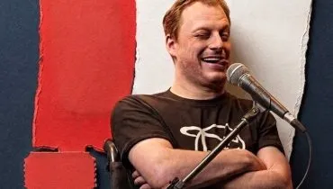 Imagem mostra um homem de braços cruzados em frente a um painel com as cores preto, vermelho e branco. Ele está fazendo uma careta e na frente de um microfone.