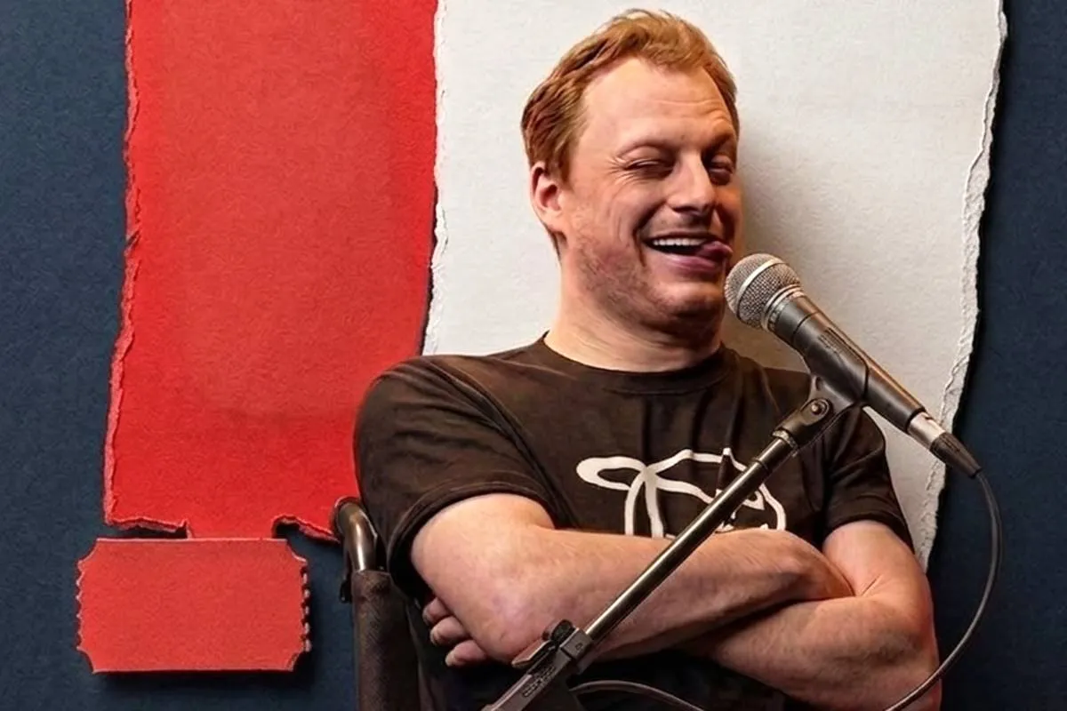 Imagem mostra um homem de braços cruzados em frente a um painel com as cores preto, vermelho e branco. Ele está fazendo uma careta e na frente de um microfone.