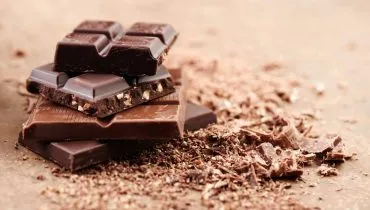 Imagem mostra quadradinhos de chocolates dispostos um em cima do outro, rom raspas em uma mesa.