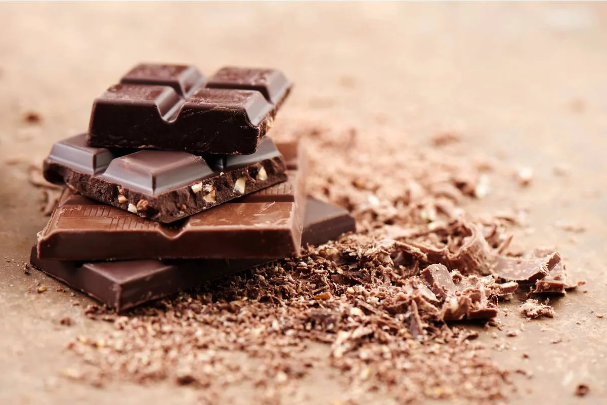 Imagem mostra quadradinhos de chocolates dispostos um em cima do outro, rom raspas em uma mesa.