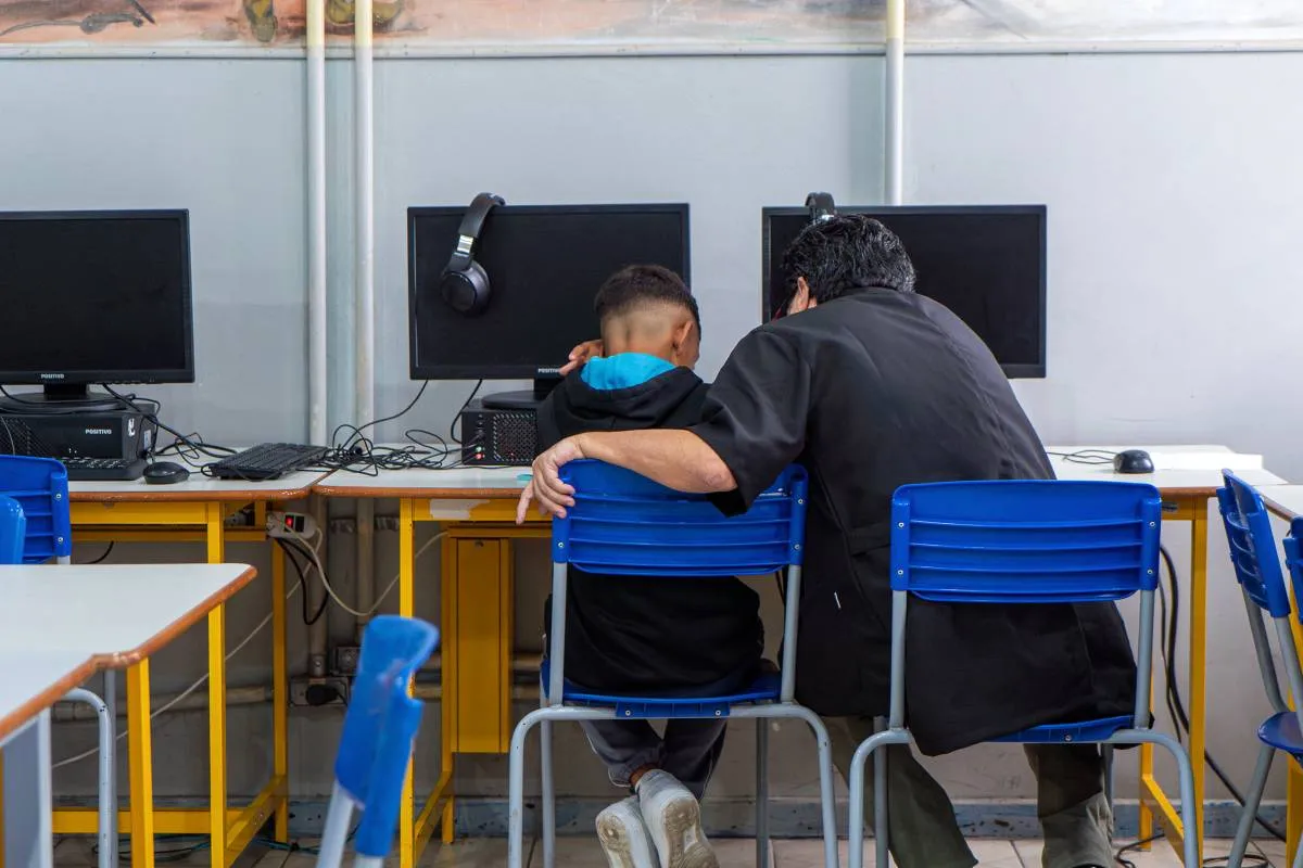 Imagem mostra um aluno com o pai lendo uma apostila na frente de um computador.