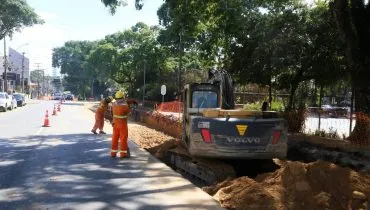 Imagem mostra uma máquina trabalhando em uma rua com uma pista bloqueada e trabalhadores da prefeitura bloqueando o trânsito.