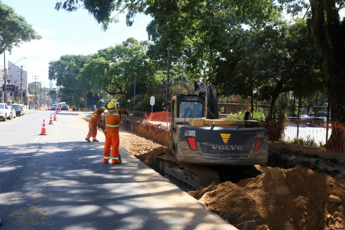Imagem mostra uma máquina trabalhando em uma rua com uma pista bloqueada e trabalhadores da prefeitura bloqueando o trânsito.