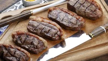 Imagem mostra um churrasco em uma tábua de madeira, com ima faca e um amolador.