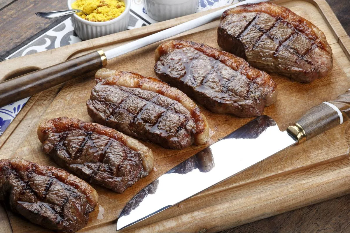 Imagem mostra um churrasco em uma tábua de madeira, com ima faca e um amolador.