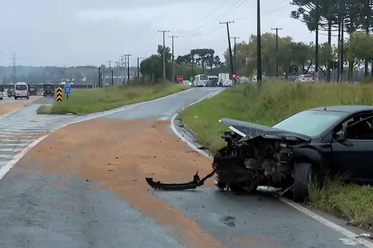 Imagem mostra um carro preto acidentado em uma curva com serragem no asfalto.