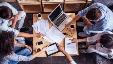 Imagem mostra pessoas trabalhando em uma mesa com notebooks e mais itens de trabalho como canetas, café e celulares.