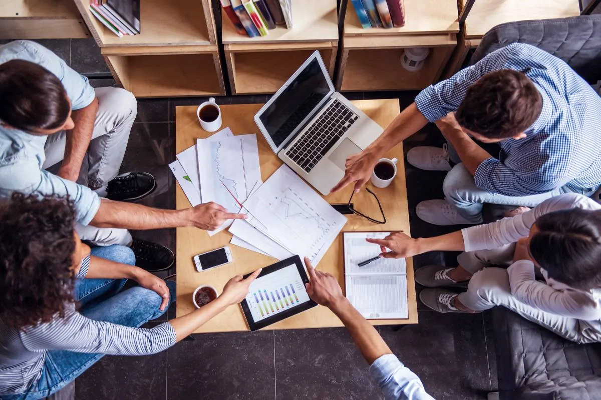 Imagem mostra pessoas trabalhando em uma mesa com notebooks e mais itens de trabalho como canetas, café e celulares.