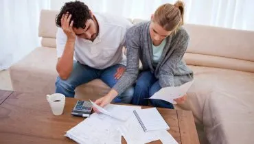 Imagem mostra um casal em uma mesa com muitas dívidas e boletos. Ele está com a mão na cabeça e a mulher segura papeis ao lado de uma calculadora.