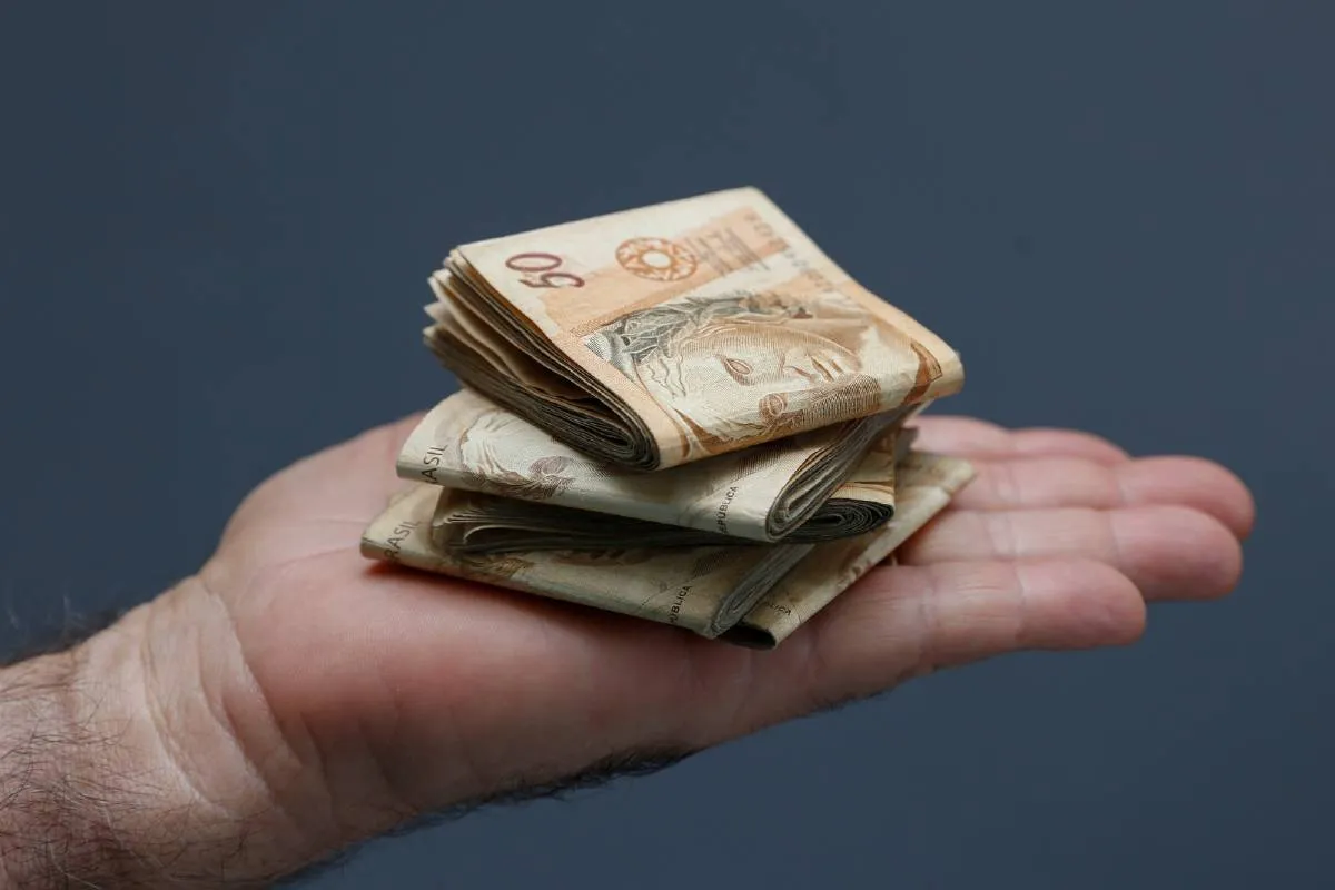 Imagem mostra um close de uma mão com varias notas de R$ 50 dobradas.