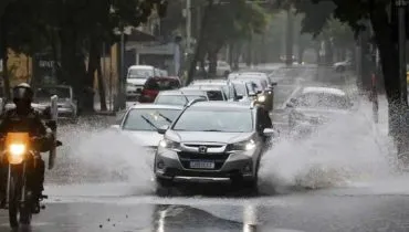 Imagem mostra carros passando em uma rua alagada por causa de um temporal.