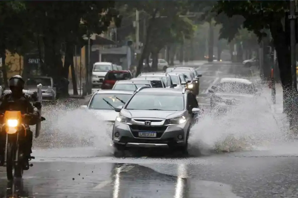 Imagem mostra carros passando em uma rua alagada por causa de um temporal.