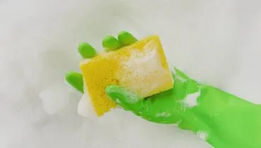 Imagem mostra uma pessoa com uma luva verde utilizando uma esponja amarela com detergente e vinagre, a mistura econômica e eficaz.