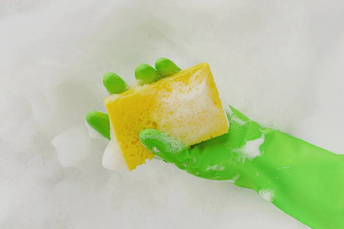 Imagem mostra uma pessoa com uma luva verde utilizando uma esponja amarela com detergente e vinagre, a mistura econômica e eficaz.