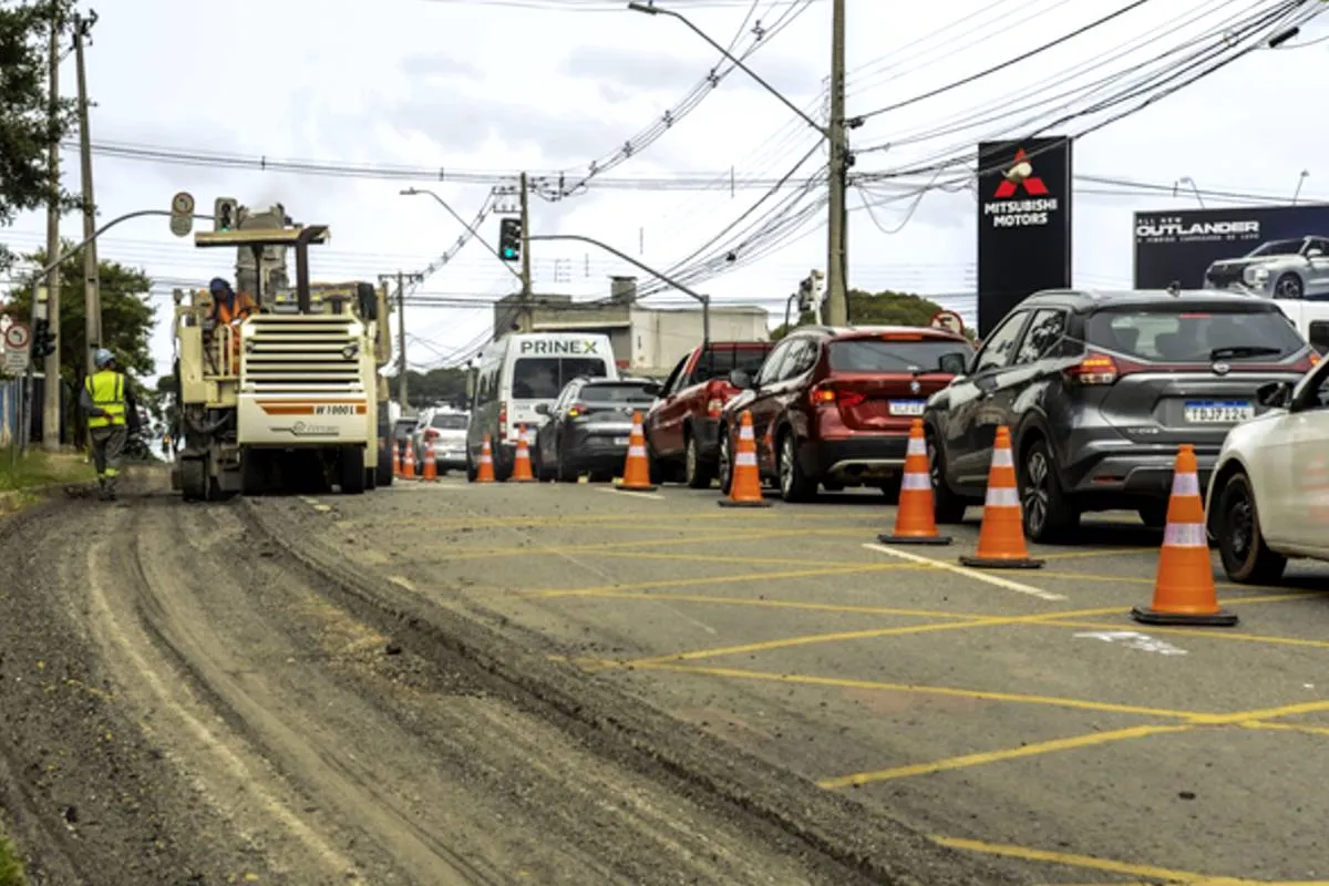 Imagem mostra uma máquina trabalhando no asfalto e uma fila grande de veículos em um engarrafamento.