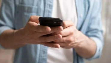 Imagem mostra um homem segurando um celular na mão. Ele veste uma camiseta branca e uma camisa azul por cima.