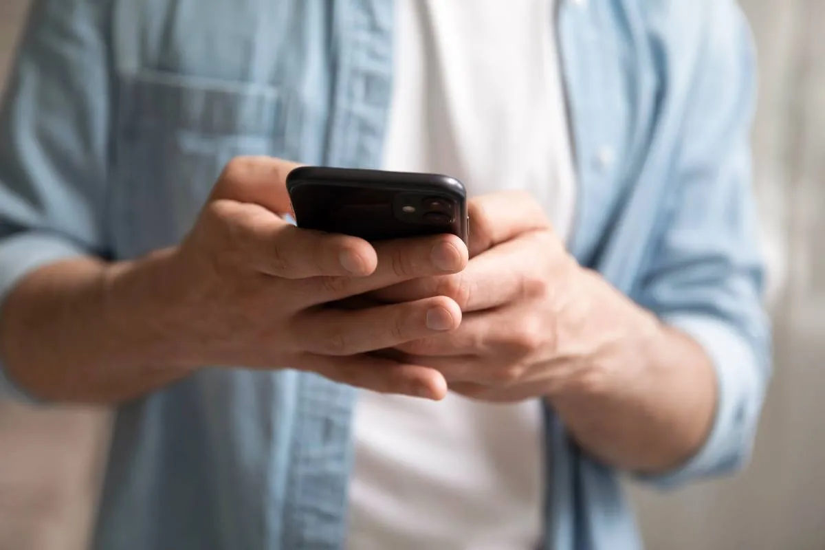 Imagem mostra um homem segurando um celular na mão. Ele veste uma camiseta branca e uma camisa azul por cima.