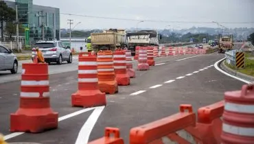 Imagem mostra cones de trânsito em um trecho da Linha Verde, em um dia nublado e com caminhões e carros circulando ao fundo.