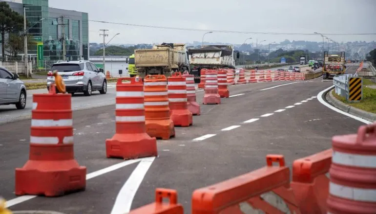 Imagem mostra cones de trânsito em um trecho da Linha Verde, em um dia nublado e com caminhões e carros circulando ao fundo.