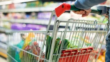 Imagem mostra uma pessoa em um supermercado empurrando um carrinho com detalhes em vermelho carregado com furtas e verduras. Ao fundo, uma prateleira com produtos.