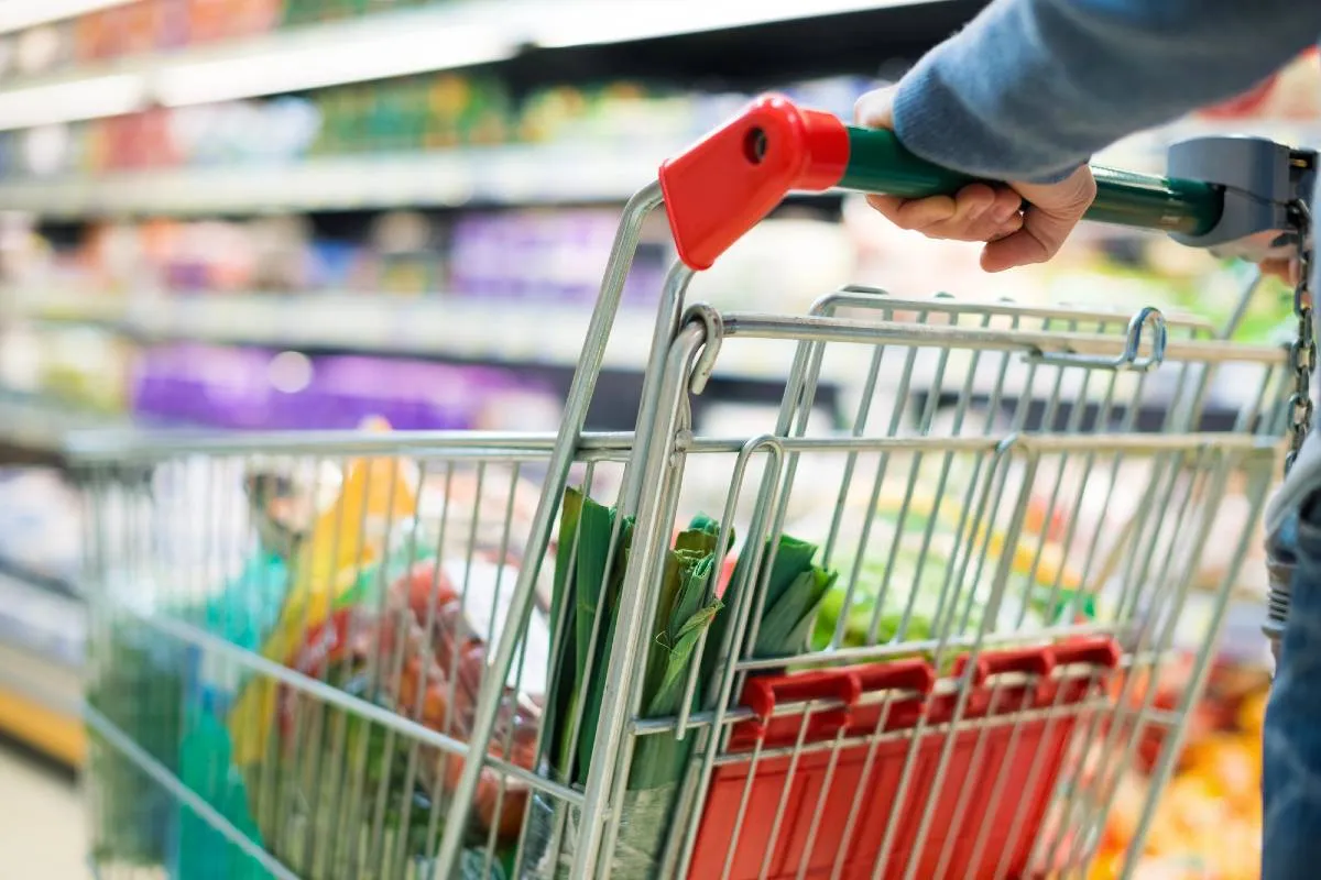 Imagem mostra uma pessoa em um supermercado empurrando um carrinho com detalhes em vermelho carregado com furtas e verduras. Ao fundo, uma prateleira com produtos.