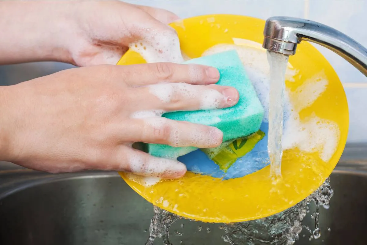Imagem mostra uma pessoa limpando um prato amarelo com uma esponja azul debaixo da torneira.
