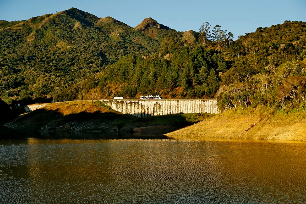 Imagem mostra parte da represa do Capivari, no nível da água, com a BR-116 ao fundo, com caminhão passando pela estrada