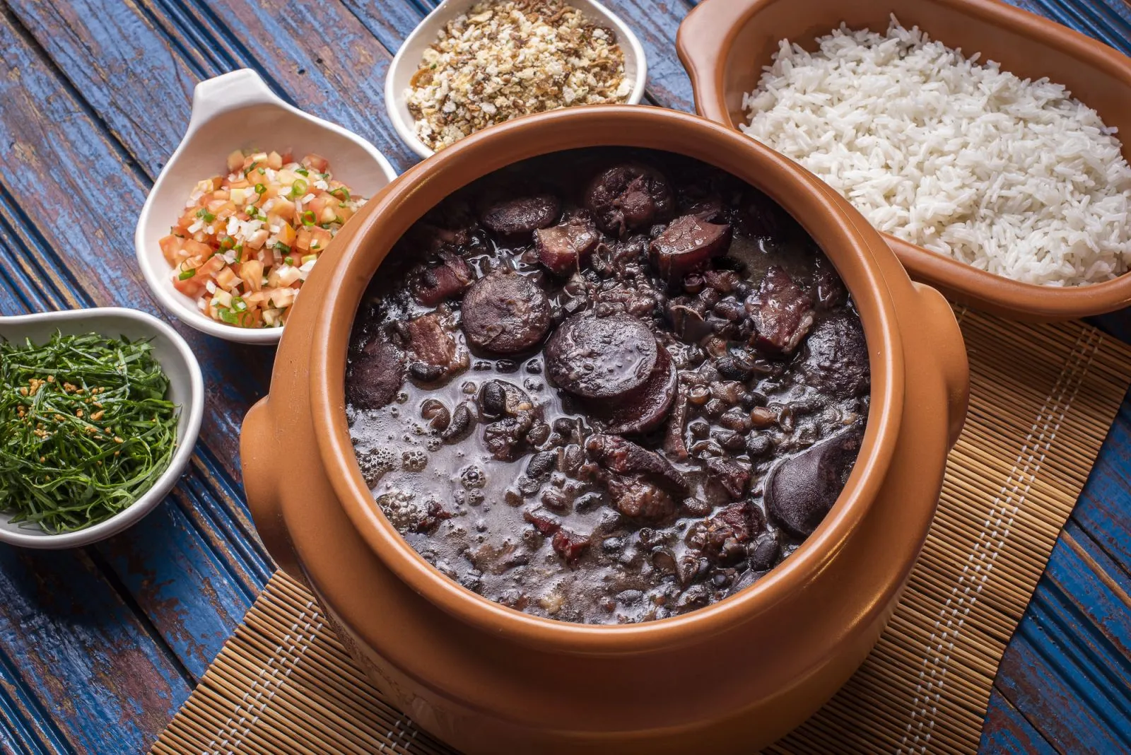 imagem mostra panela de feijoada, com farofa, couve, vinagrete e arroz de acompnhamento