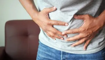 Imagem mostra um homem com as mãos na barriga num momento de constipação. Ele usa camisa cinza e calça jeans.
