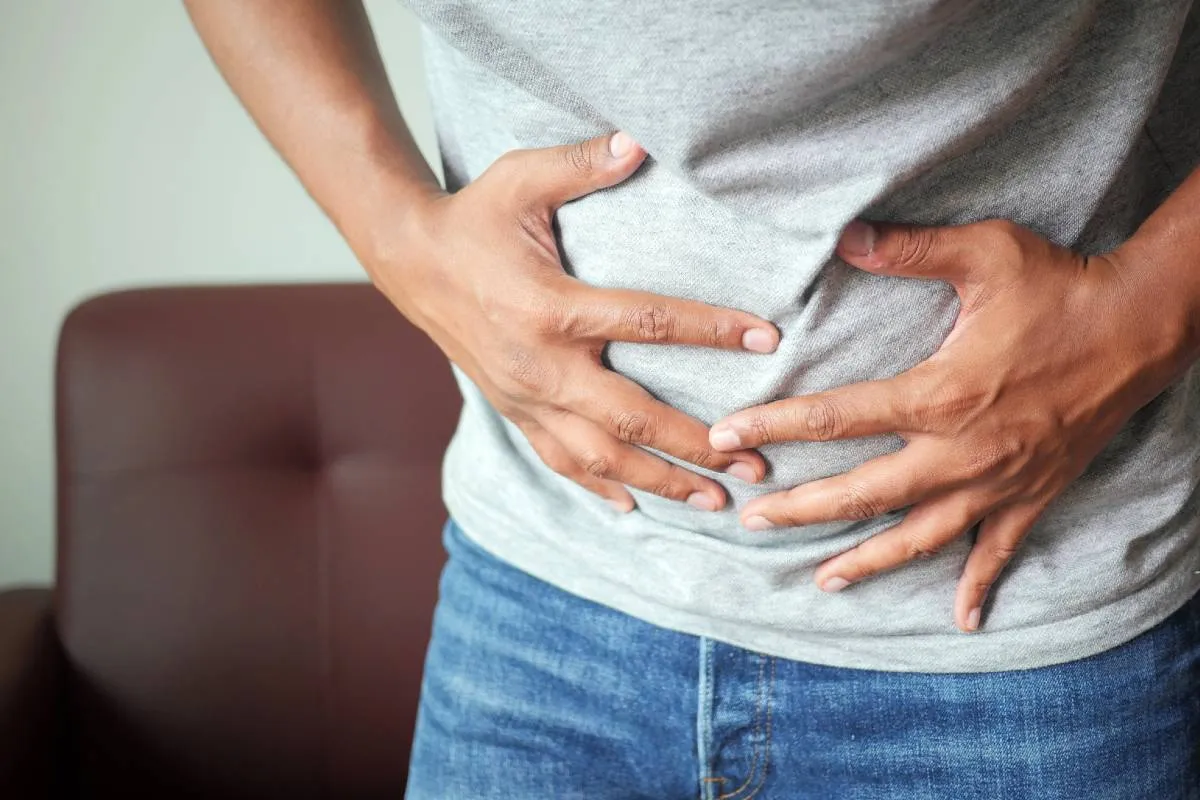 Imagem mostra um homem com as mãos na barriga num momento de constipação. Ele usa camisa cinza e calça jeans.
