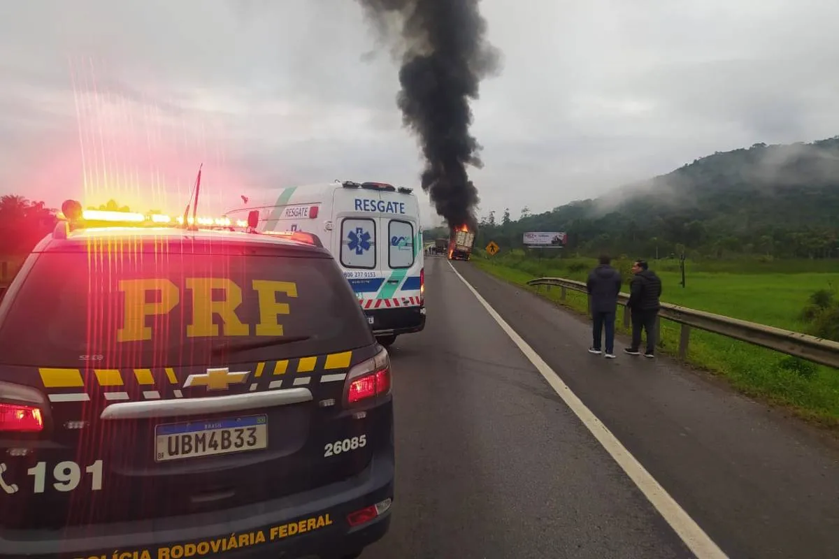 Imagem mostra uma viatura da PRF parada perto de um caminhão em chamas. Hpa uma grande coluna de fumaça preta ao fundo da imagem e uma ambulância branca no meio.