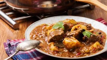 Imagem mostra um prato com vaca atolada feita na panela de pressão com mandioca. O prato está vem servido, com uma colhar de prata apoiada em uma mesa de madeira com um pano azul em baixo.