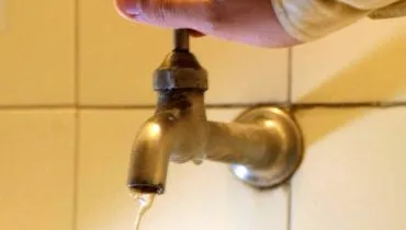 Imagem mostra uma torneira com pouca água saindo e uma pessoa tentando abrir mais. Ao fundo, azulejos.