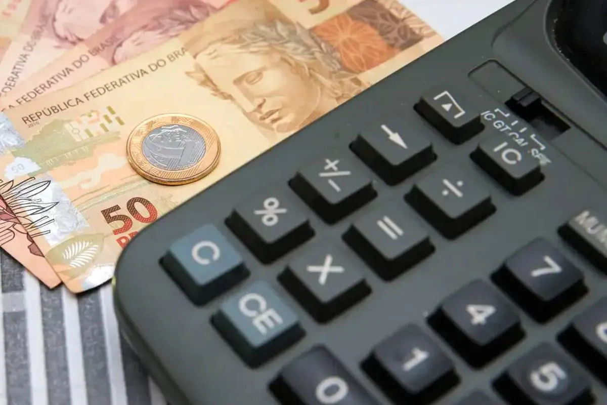Imagem mostra uma calculadora e notas de real com moedas.