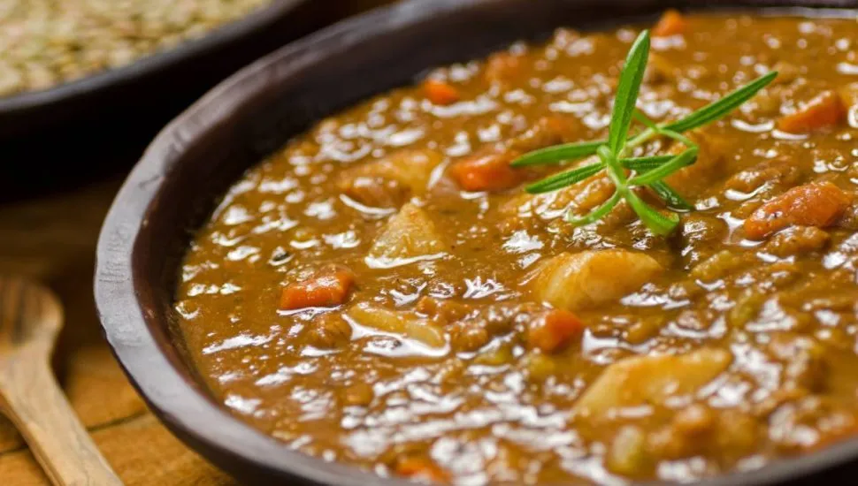 Imagem mostra um prato com uma deliciosa sopa de legumes. Tem uma colher de pau ao lado e um ramo de alecrim em cima.