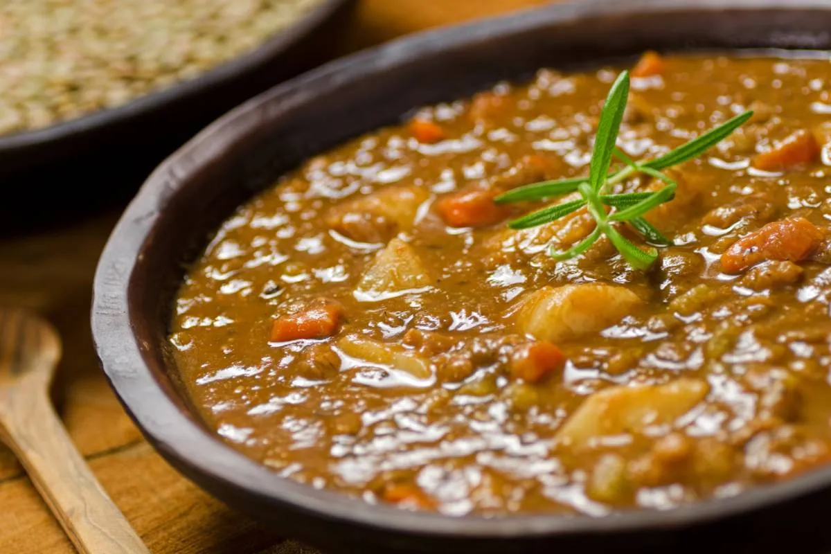 Imagem mostra um prato com uma deliciosa sopa de legumes. Tem uma colher de pau ao lado e um ramo de alecrim em cima.