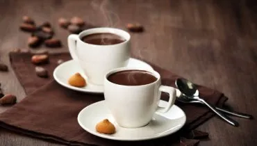 Duas xícaras brancas com chocolate quente sobre uma mesa de madeira escura, com leve fumaça saindo da bebida. A cena tem decoração com grãos de cacau, pedaços de chocolate e biscoitos, em clima aconchegante de inverno.