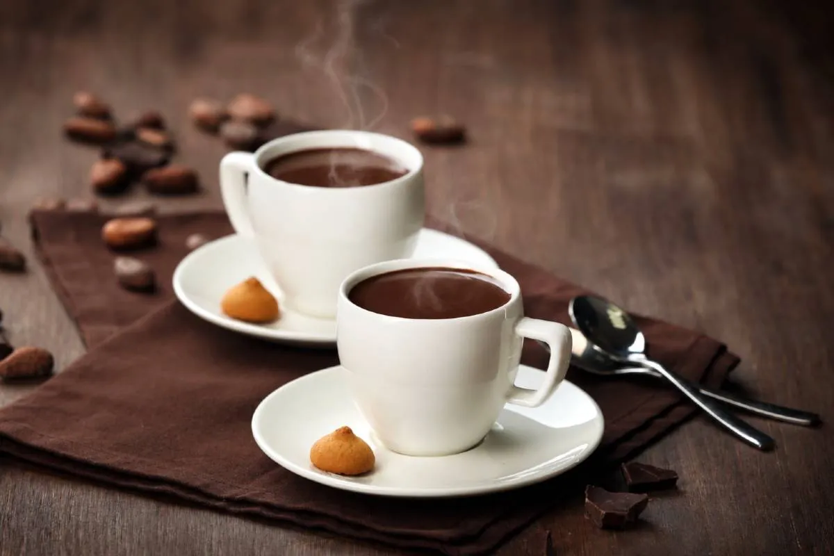 Duas xícaras brancas com chocolate quente sobre uma mesa de madeira escura, com leve fumaça saindo da bebida. A cena tem decoração com grãos de cacau, pedaços de chocolate e biscoitos, em clima aconchegante de inverno.