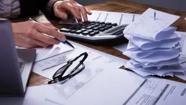 Imagem mostra uma pessoa trabalhando em uma mesa com muitos papeis, uma calculadora, um óculos e um computador.