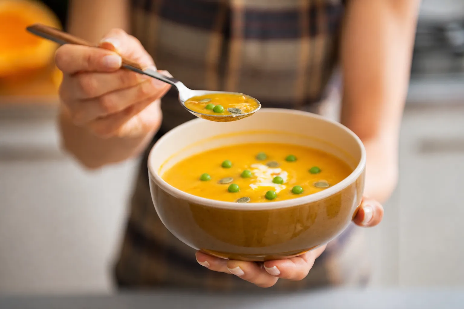 Imagem mostra uma pessoa segurando uma tigela de sopa cremosa alaranjada enquanto leva uma colher à boca. A sopa está decorada com ervilhas, sementes e um toque de creme, em um ambiente aconchegante de cozinha.